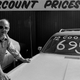 A black and white photo of a man leaning on a used car at a dealership.